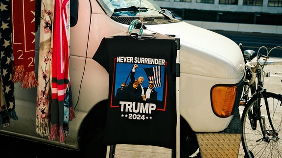 An der Radioantenne eines weißen Trucks hängt ein schwarzes T-Shirt, auf dem Donald Trump seine Faust in die Höhe reckt, während ihn drei Männer mit Sonnenbrille abschirmen.