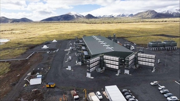 Luftbild des weltweit größten CO2-Direktabscheiders, der auf Island in Betrieb gegangen ist, eine Industriehalle mit mehreren Vorbauten, im Hintergrund die Berge von Island.