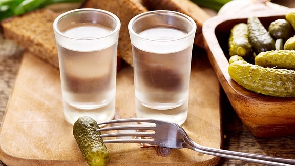 Zwei kleine Gläser Wodka mit sauren Gurken und Brotscheiben auf einem Holztisch.