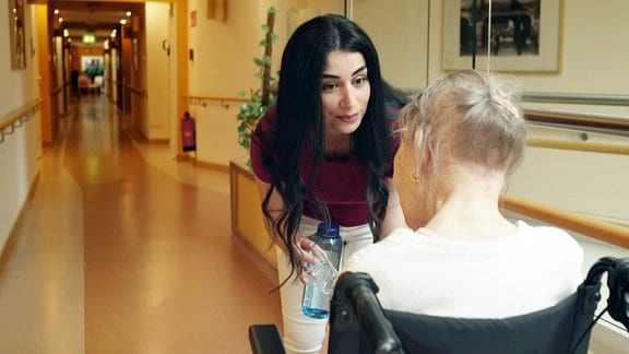 Eine junge Pflegerin beugt sich zu einer Patientin im Rollstuhl runter. 