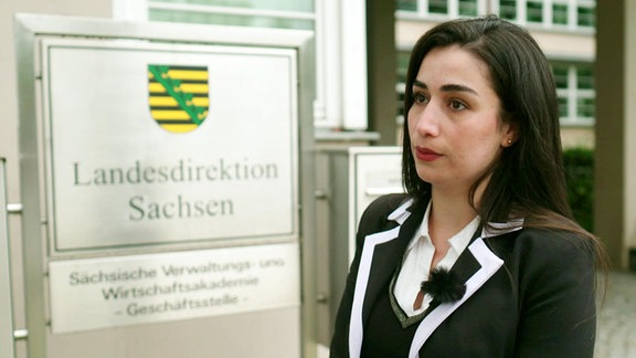 Eine Frau mit schwarzen Haaren vor dem Schild mit der Aufschrift "Landesdirektion Sachsen".