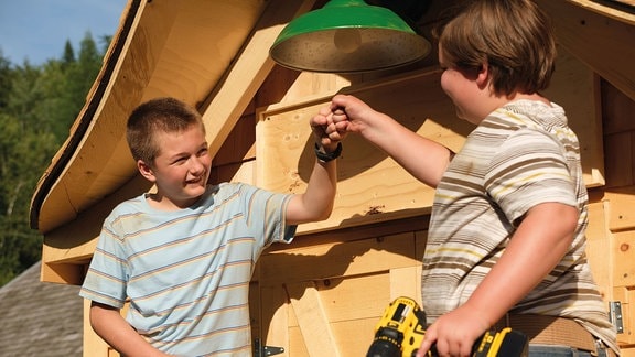 Zwei Jungs mit Akkuschraubern in der Hand freuen sich vor einer Holzhütte.