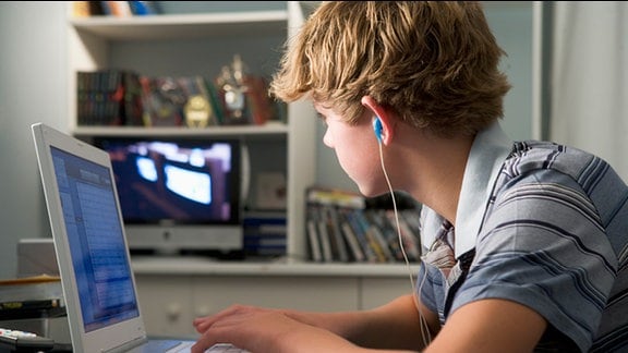Jugendlicher, mit Kopfhörern im Ohr, sitzt vor einem Laptop und schaut auf einen Ferseher