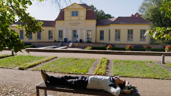 Ein Mann liegt auf einer Bank in einem Park, im Hintergrund ein Gebäude mit gelber Fassade. 