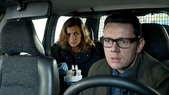 Szene aus Tatort Dresden, ein Mann mit dunkler Brille sitzt am Lenkrad eines Autos, auf dem Rücksitz eine Frau
