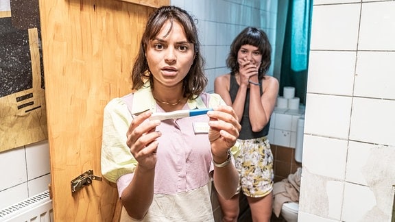 Paula (Taneshia Abt) steht mit einem Schwangerschaftstest in der Hand in einer Klotür. Hinter ihr steht ihre Freundin Kim (Johanna Franke).