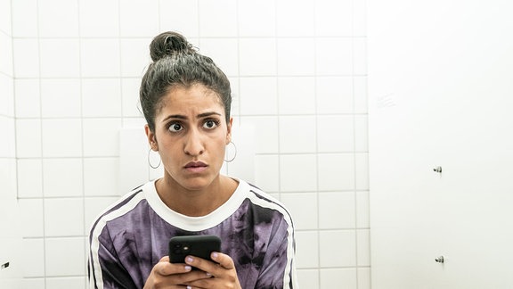 Samira (Banafshe Hourmazdi) sitzt mit dem Handy in der Hand auf einer Toilette.