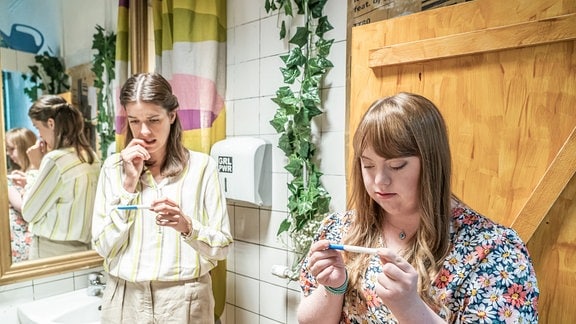 Neele (Marie Nasemann) und ihre Cousine Tina (Louisa Wöllisch) stehen mit im Vorraum zu einer Toilette. Beide haben einen Schwangerschaftstest in der Hand und schauen gespannt darauf.