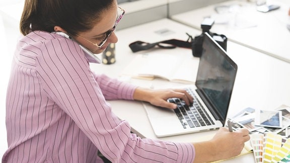 Junge Frau telefoniert während sie gleichzeitig am Laptop arbeitet und etwas notiert.