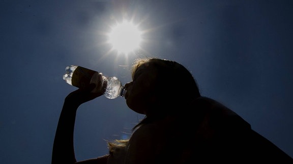 Eine Frau trinkt vor einer strahlenden Sonne aus einer Wasserflasche.