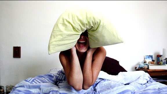 Eine Frau sitzt schreiend im Bett und hält sich mit dem Kopfkissen die Ohren zu