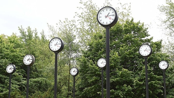 Installation aus mehreren Uhren, die auf Metallsäulen stehen und alle die gleiche Zeit anzeigen.