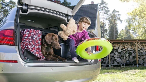 Ein kleines Mädchen spielt in einem beladenen Pkw-Kofferraum neben einem Hund.  