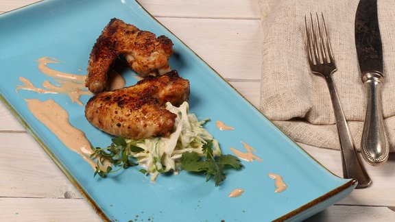 Auf einem blauen Teller liegen zwei Chicken Wings und feiner Salat, garniert mit einem hellen Dip.