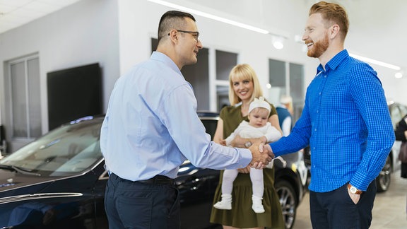 Ein Ehepaar mit Kind bei einem Autohändler. Die Männer geben sich die Hand.