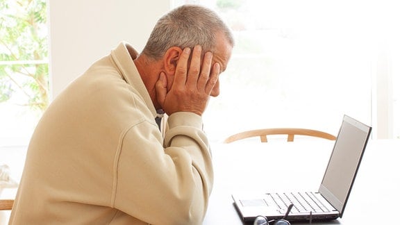 Ein alter Mann sitzt am Computer und schaut verzweifelt auf den Bildschirm. Er stützt den Kopf in die Hände.