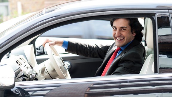 Ein Mann im Anzug sitzt hinter dem Lenkrad eines Autos und lacht in die Kamera