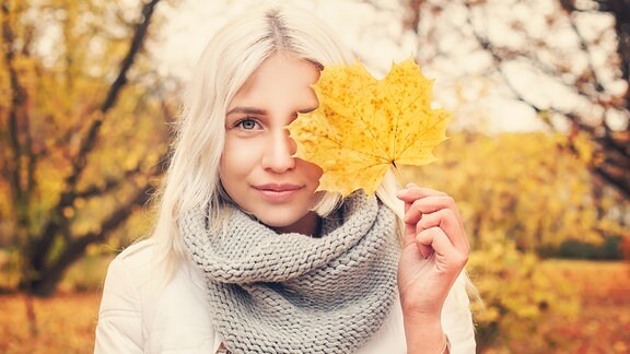 Eine hübsche blonde Frau hält sich ein Herbstblatt vor ihr linkes Auge.