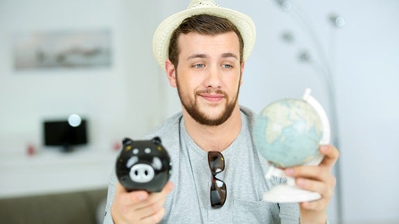 Ein hübscher Mann mit Strohhut hält einen kleinen Globus und ein Sparschwein in seinen Händen