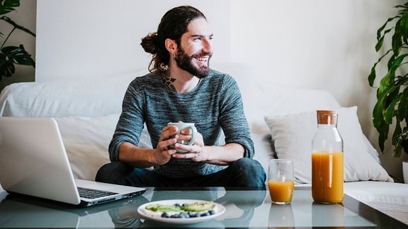 Lächelnder Mann mit Kaffeetasse, der weg schaut, während des Frühstücks auf dem Sofa zu Hause sitzt