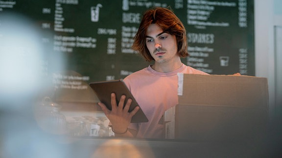 Ernst blickender Ladenbesitzer hat eine Kiste vor sich auf dem Tresen und einen Tablet-PC in der hand