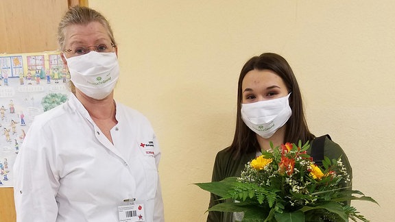 Zwei Frauen mit Mund-Nasen-Schutz stehen nebeneinander, eine hält einen Blumenstrauß