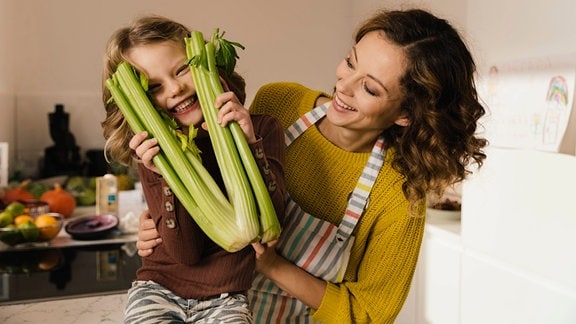 Mädchen mit Mutter in einer Küche schaut lachend durch Selleriestaude