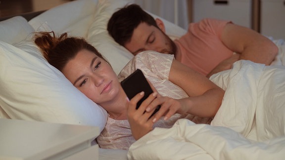 Ein Pärchen liegt im Bett. Der Mann schläft und die Frau spielt am Handy.