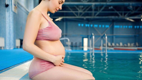 Eine schwangere Frau sitzt am Beckenrand eines Schwimmbades.