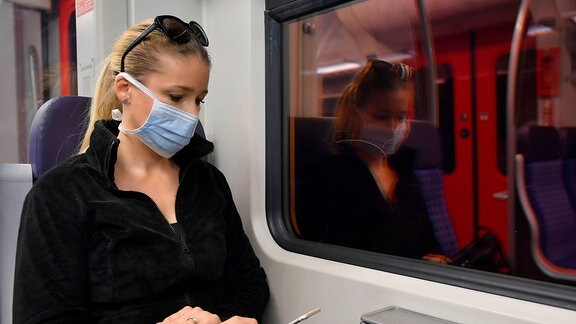 Eine junge Frau sitzt mit Smartphone und Mundschutz im Zug.