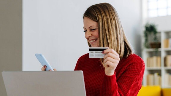 Fröhliche Frau vor einem Laptop mit Smartphone und Kreditkarte in den Händen