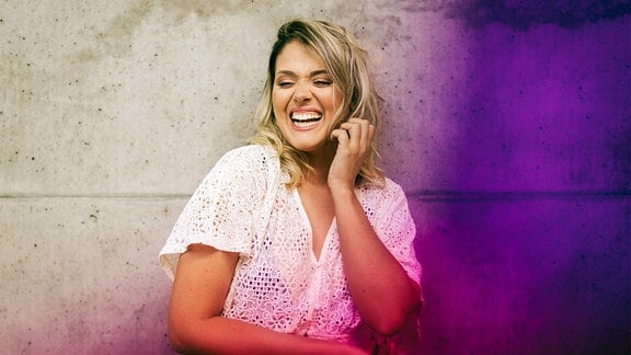Lachende Frau steht im Freien an lilafarbener Wand.