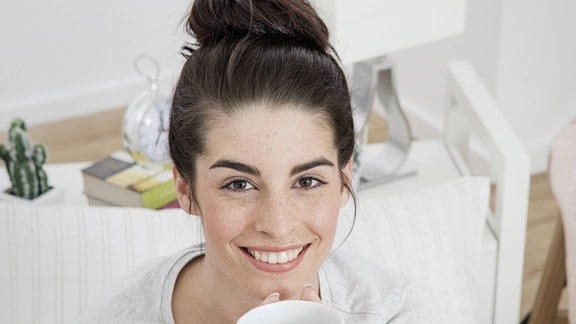 Junge Frau mit hochgeteckten Haaren sitzt mit Kaffeetasse im Bett.