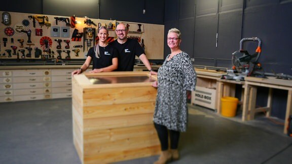 Drei Menschen stehen neben einem Hochbeet aus Holz in einer Werkstatt.