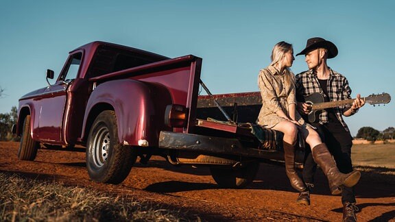 Junger Mann mit Cowboyhut und Gitarre sitzt neben einer jungen Frau auf der Ladefläche eines Retro-Pickup-Autos.