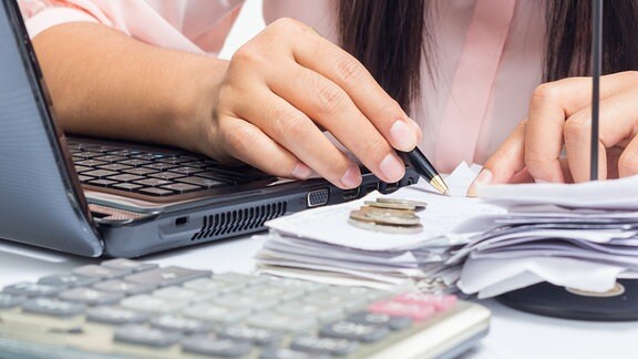 Frau sitzt mit Stift, Rechnungen, Münzen, Taschenrechner und Laptop am Tisch