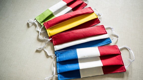 Selbstgenähte Atemschutzmasken in den Farben der Flaggen von Italien, Frankreich, Niederlande und Spanien liegen auf einem Tisch