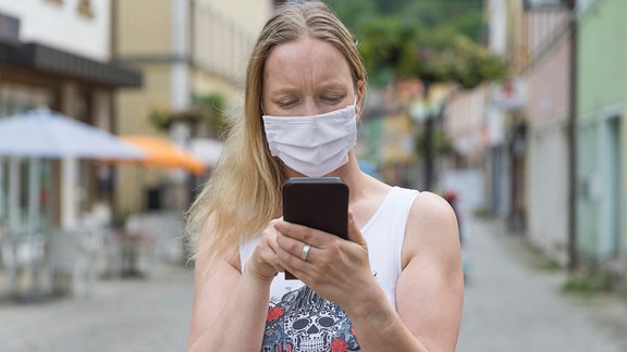 Eine junge Frau mit Mund-Nasen-Behelfs-Schutz schaut in einer Fußgängerzone auf ihr Handy. 