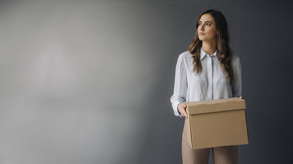 Eine ernst blickende, junge Businessfrau hält einen Karton in ihren Händen, während sie vor einer grauen Wand steht.