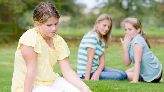 Hinter traurigem Mädchen auf einer Wiese lästern zwei weitere Mädchen.