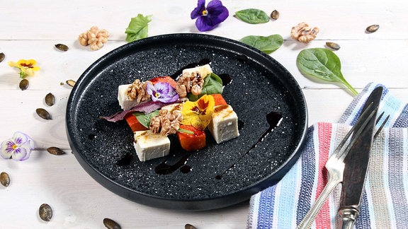 Grob geschnittete Kürbisstücke mit Walnüssen und Blüten auf einem dunklen Teller.