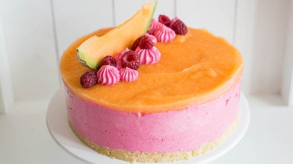 Eine kleine Torte in rosa und orange, dekoriert mit Himbeeren und einer Melonenscheibe.