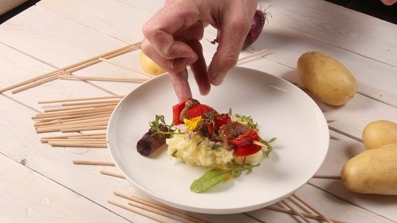 Eine Hand greift nach einem Schaschlik-Spieß, der auf Kartoffelbrei auf einem dekorierten Teller liegt.