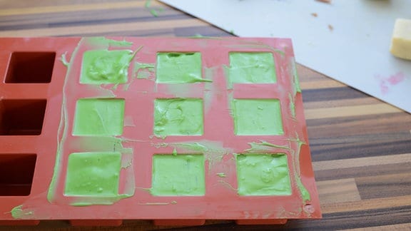 Eine Backform aus Silikon mit grüner Füllung neben vielen kleinen quadratischen Küchlein