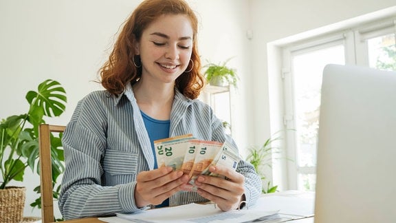 Eine junge Frau am Laptop lächelt und hat Geldscheine in der Hand
