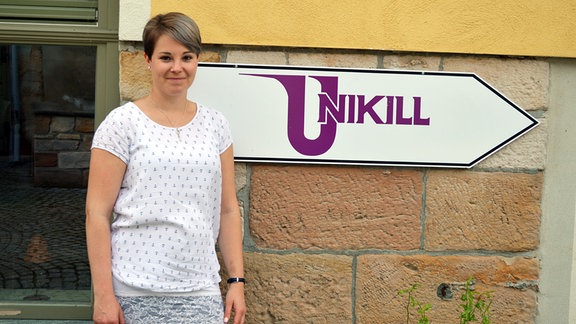 Eine junge Frau steht vor einem Schild mit der Aufschrift: Unikill.