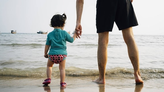 Kind mit UV-Schutz-T-Shirt an der Hand seines Vaters am Strand