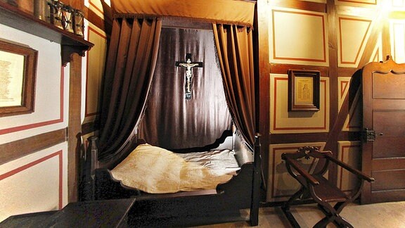 Nachbau von Luthers Schlafzimmer mit seinem Bett, vor dem lange Vorhänge seitlich gerafft sind. Daneben steht ein sehr alter Stuhl