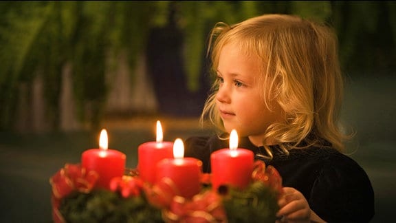 Ein blondes Mädchen neben einem Adventskranz, auf dem vier Kerzen brennen