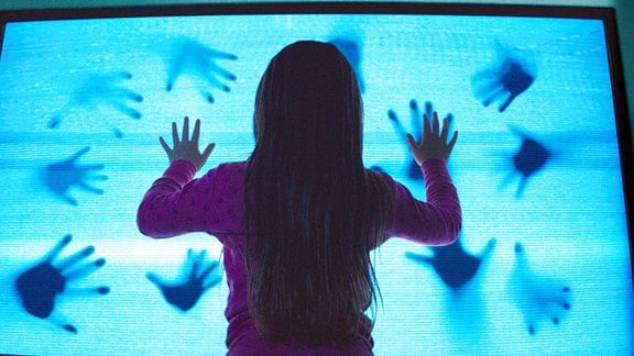 Ein Mädchen schaut auf eine blaue, leuchtende Fläche auf der von der Gegenseite Schatten von Händen zu sehen sind.